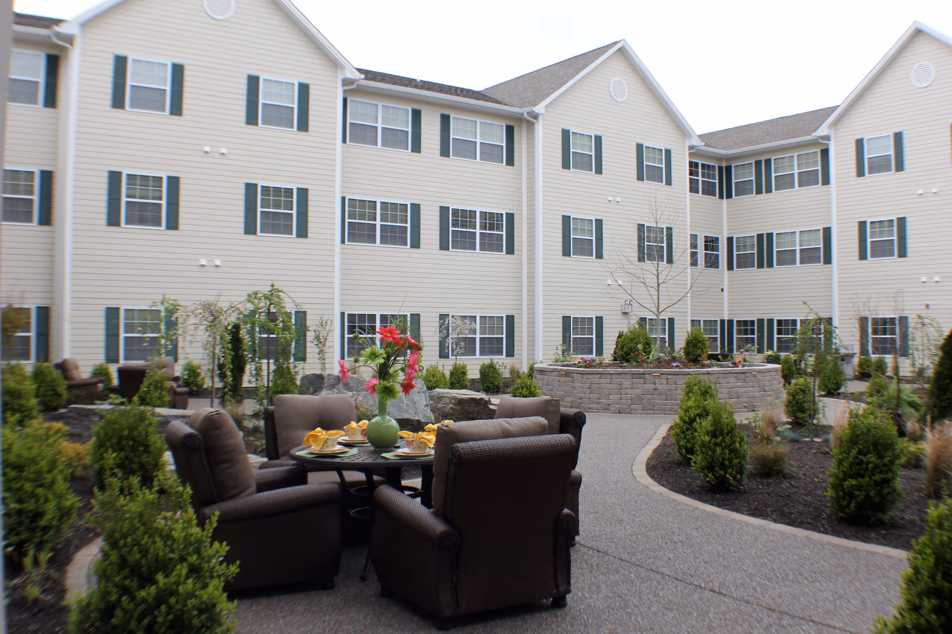 Outdoor courtyard area of a senior living facility with beige multi-story building in the background. The courtyard features a round table with four cushioned chairs, a green vase with red flowers on the table, and place settings with yellow napkins. The area is landscaped with small bushes, plants, and a raised stone planter in the center.