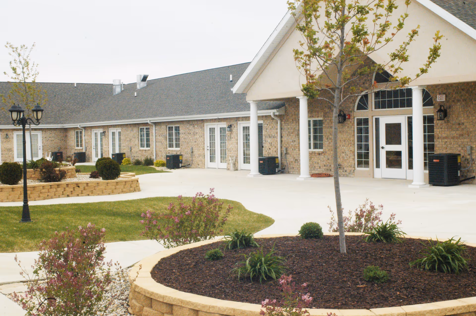 Front exterior of a single-story brick senior living facility with a covered entrance, white columns, and landscaped courtyard.