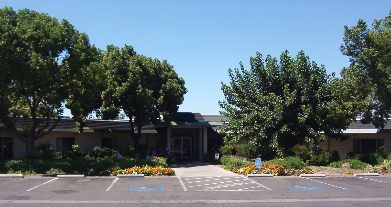 Front exterior view of Franciscan Post-Acute Care Center with a single-story building surrounded by trees and flower beds, and a parking lot with marked handicap spaces in front.