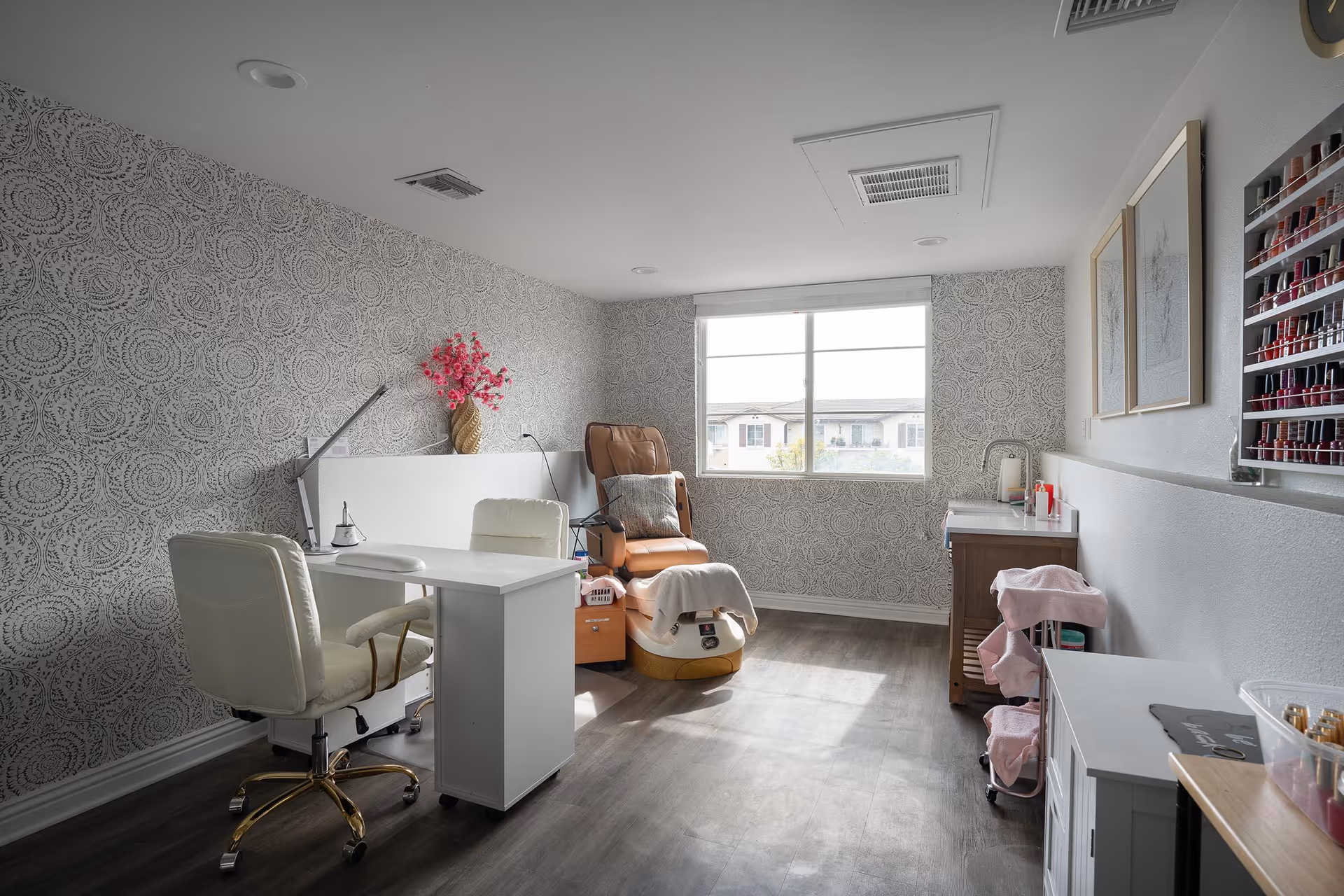 A bright room with patterned wallpaper and a large window letting in natural light. The room features a white desk with a white swivel chair, a brown leather pedicure chair with a foot bath, and a nail polish display rack on the wall. There are also some framed pictures and a vase with pink flowers on a cabinet.
