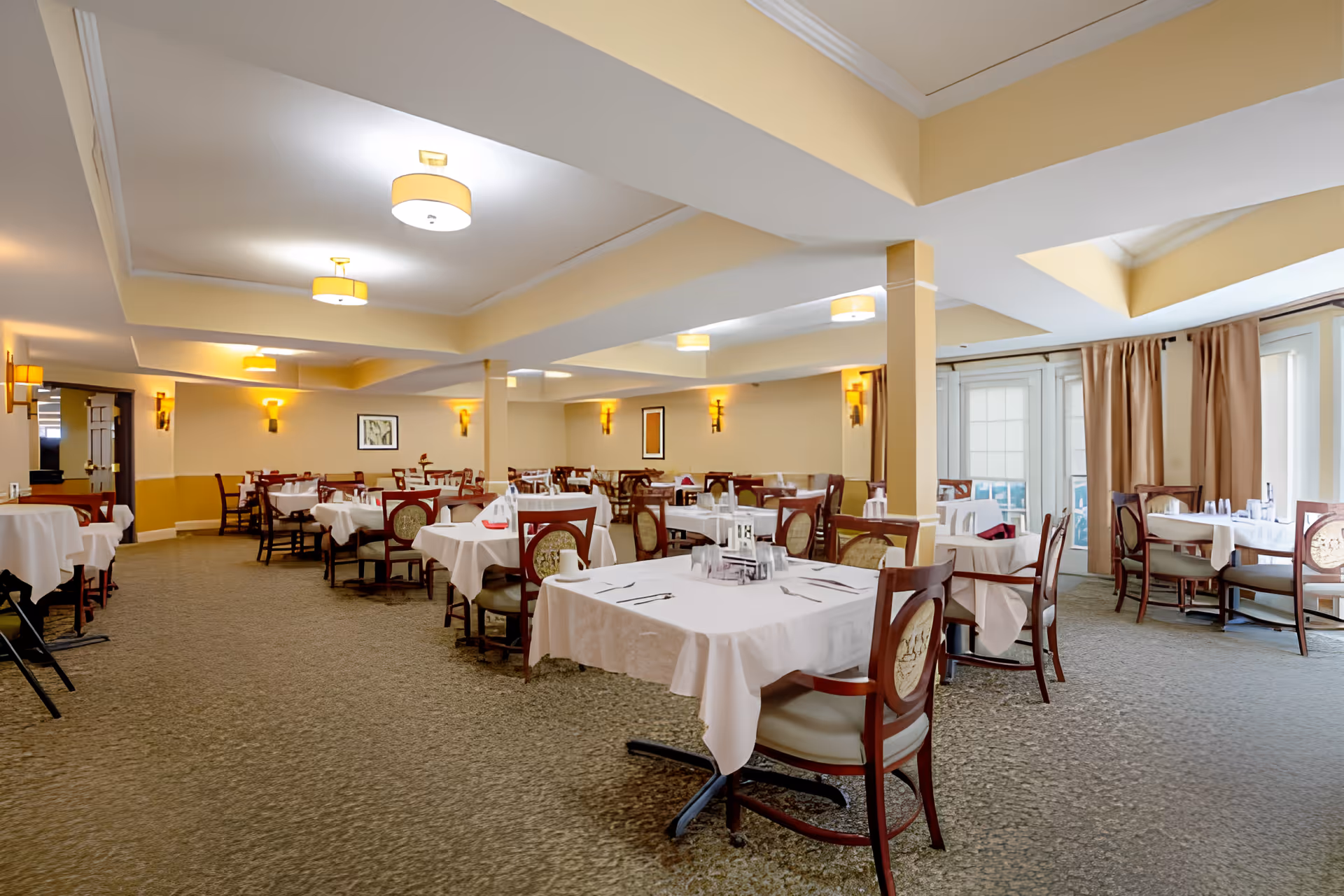 Spacious dining room with tables covered in white tablecloths and wooden chairs arranged under soft ceiling lights.