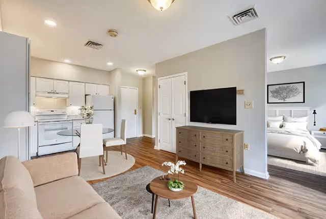 A bright and modern senior living apartment interior featuring a beige sofa, a small round wooden coffee table with a plant, a wall-mounted flat-screen TV above a wooden dresser, a round glass dining table with two white chairs, and a compact kitchen with white cabinets and appliances. A bedroom with a neatly made bed and framed artwork is visible through an open doorway.