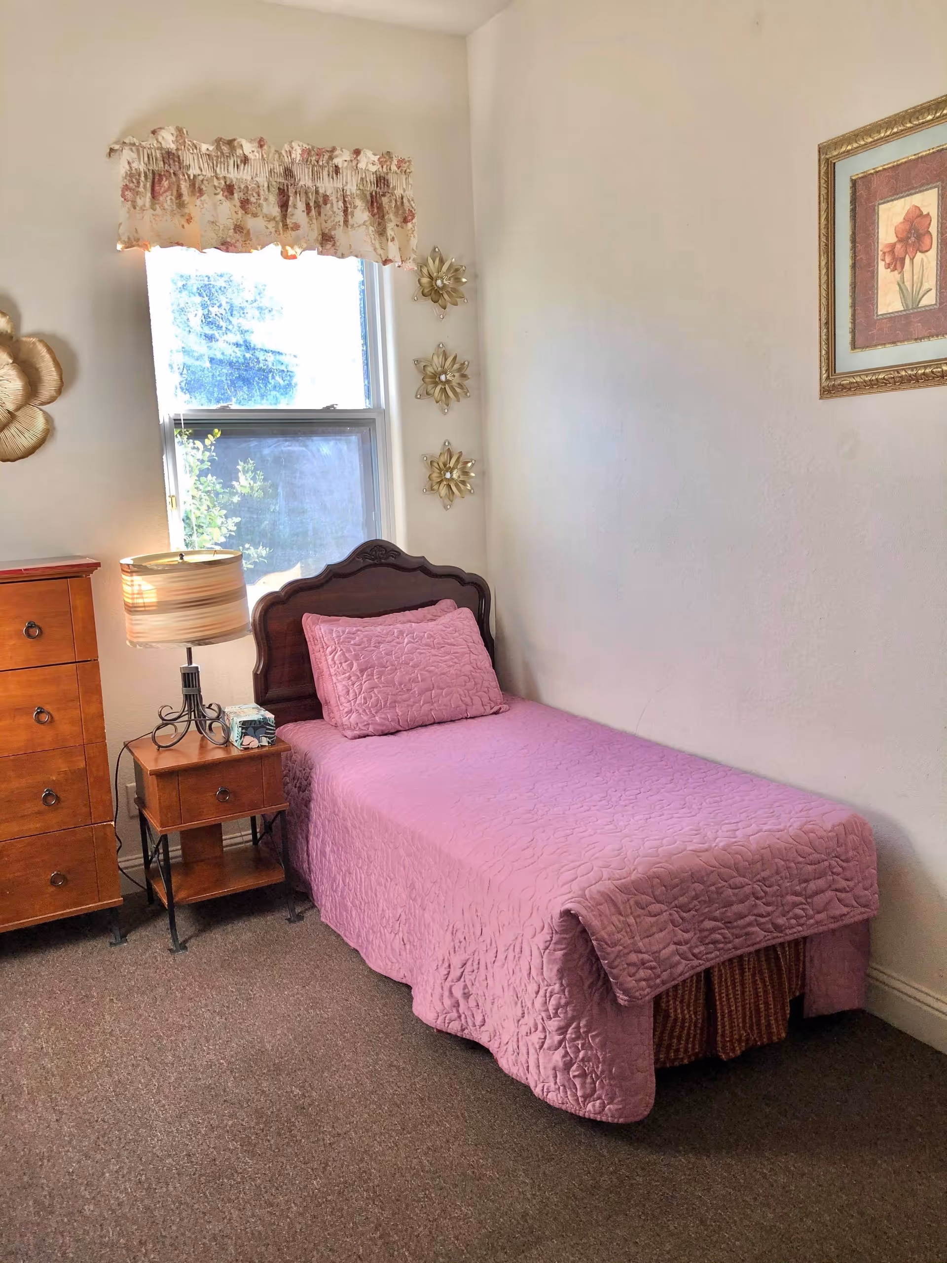 Small bedroom with a single bed covered in a pink quilt, wooden headboard, nightstand with a lamp, dresser, and a window with a floral valance.