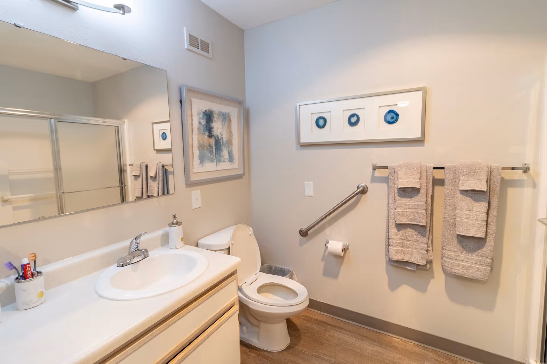 A clean and well-lit bathroom featuring a white sink with a silver faucet, a large mirror above the sink, a toilet with the lid open, two framed abstract artworks on the walls, a metal grab bar next to the toilet, a toilet paper holder, and a towel rack with three beige towels hanging on it. The floor has a wood-like finish.