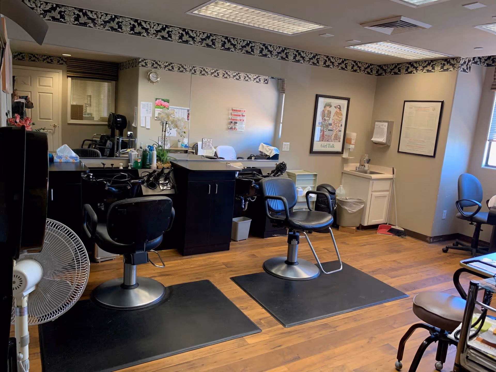 Interior view of a hair salon area in a senior living facility with two black salon chairs on mats in front of a large mirror, a fan, a small sink, and framed pictures on the wall. The room has wood flooring and fluorescent ceiling lights.
