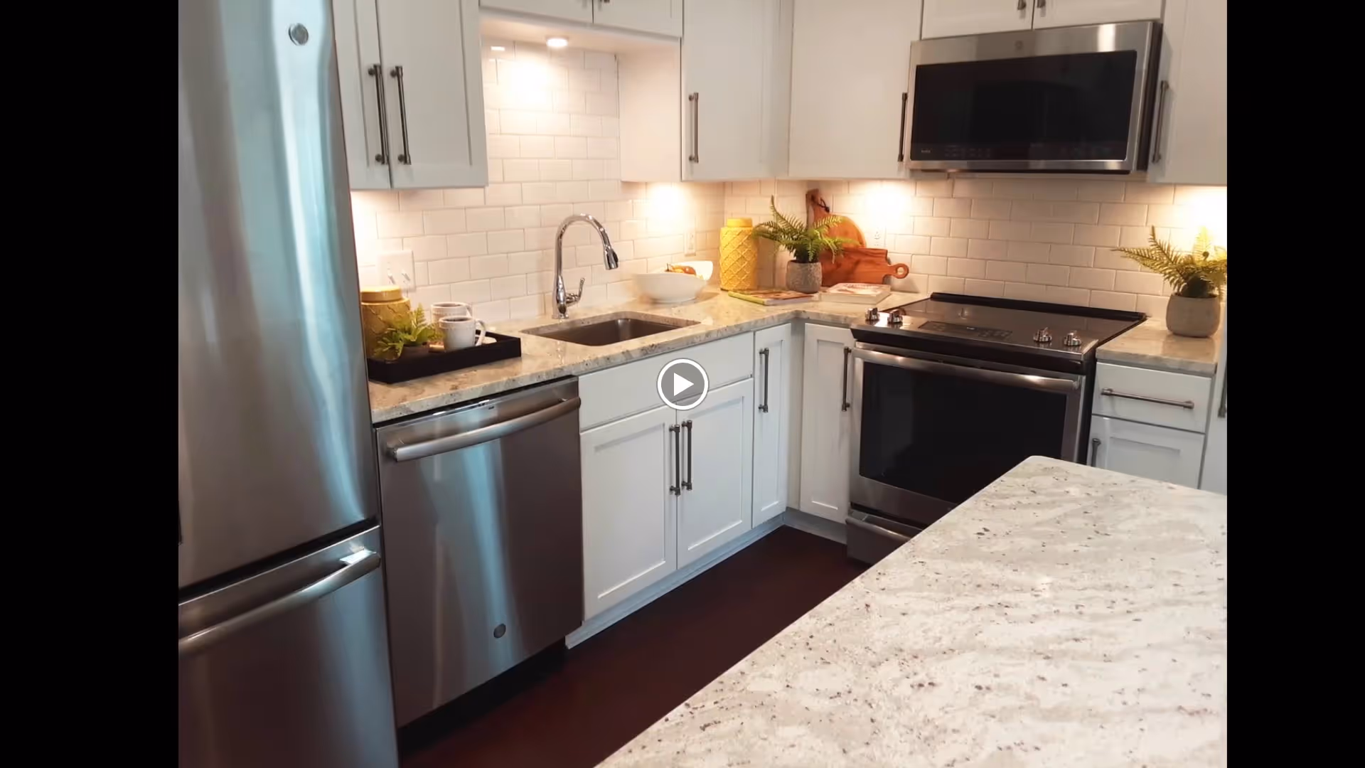 Modern kitchen with stainless steel refrigerator, dishwasher, oven, and microwave. White cabinets with silver handles, white subway tile backsplash, and light-colored granite countertops. Decorative items include a small plant, a yellow container, and a wooden cutting board.