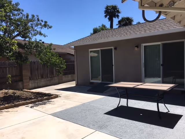 Outdoor patio area with a table on a gray outdoor rug, adjacent to a building with two sliding glass doors. There are trees and a wooden fence in the background under a clear blue sky.