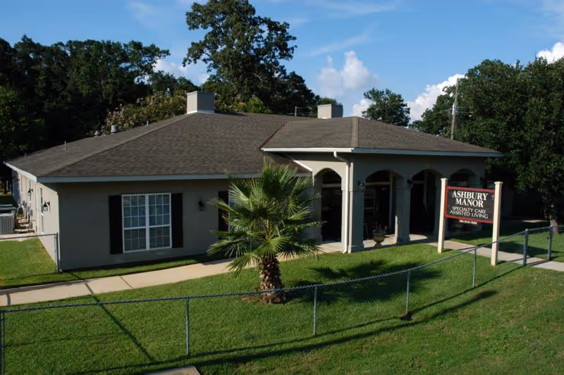 Single-story building with a gray roof and beige walls, surrounded by green grass and trees. A small palm tree is in front of the building, which has a covered porch with arches. A sign near the sidewalk reads 'Ashbury Manor Specialty Care Assisted Living.'