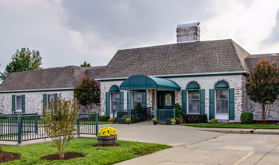 Exterior view of a single-story senior living facility building with white brick walls, green window shutters, and a green awning over the entrance. There are small trees and shrubs around the building, a paved driveway, and a flower barrel with yellow flowers near the entrance.