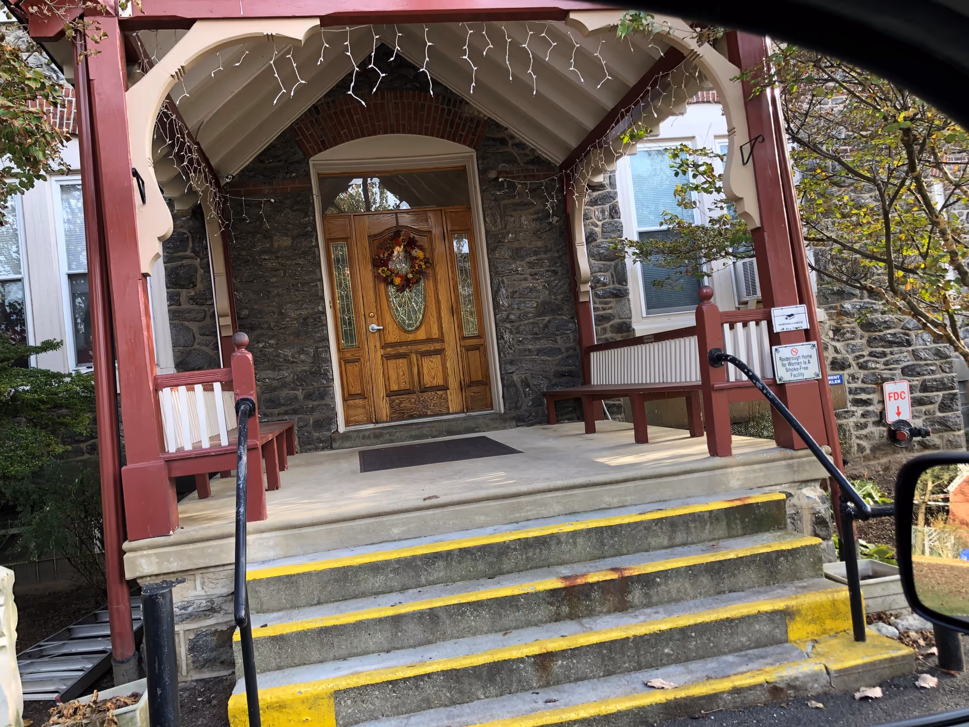 Front entrance of a stone building with a wooden door decorated with a fall wreath. The entrance has a covered porch with red and white benches on either side and steps with yellow safety markings leading up to it. There are some small trees and bushes around the porch area.