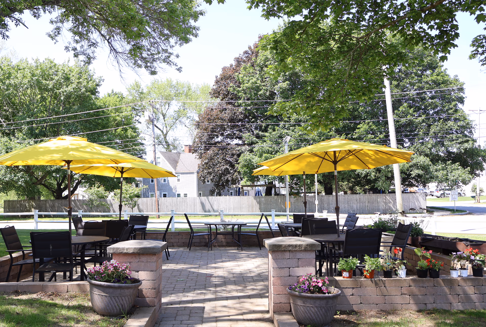 Outdoor patio area with several black tables and chairs under large yellow umbrellas. The patio is surrounded by low brick walls with potted flowers and plants. Trees and a residential neighborhood are visible in the background.