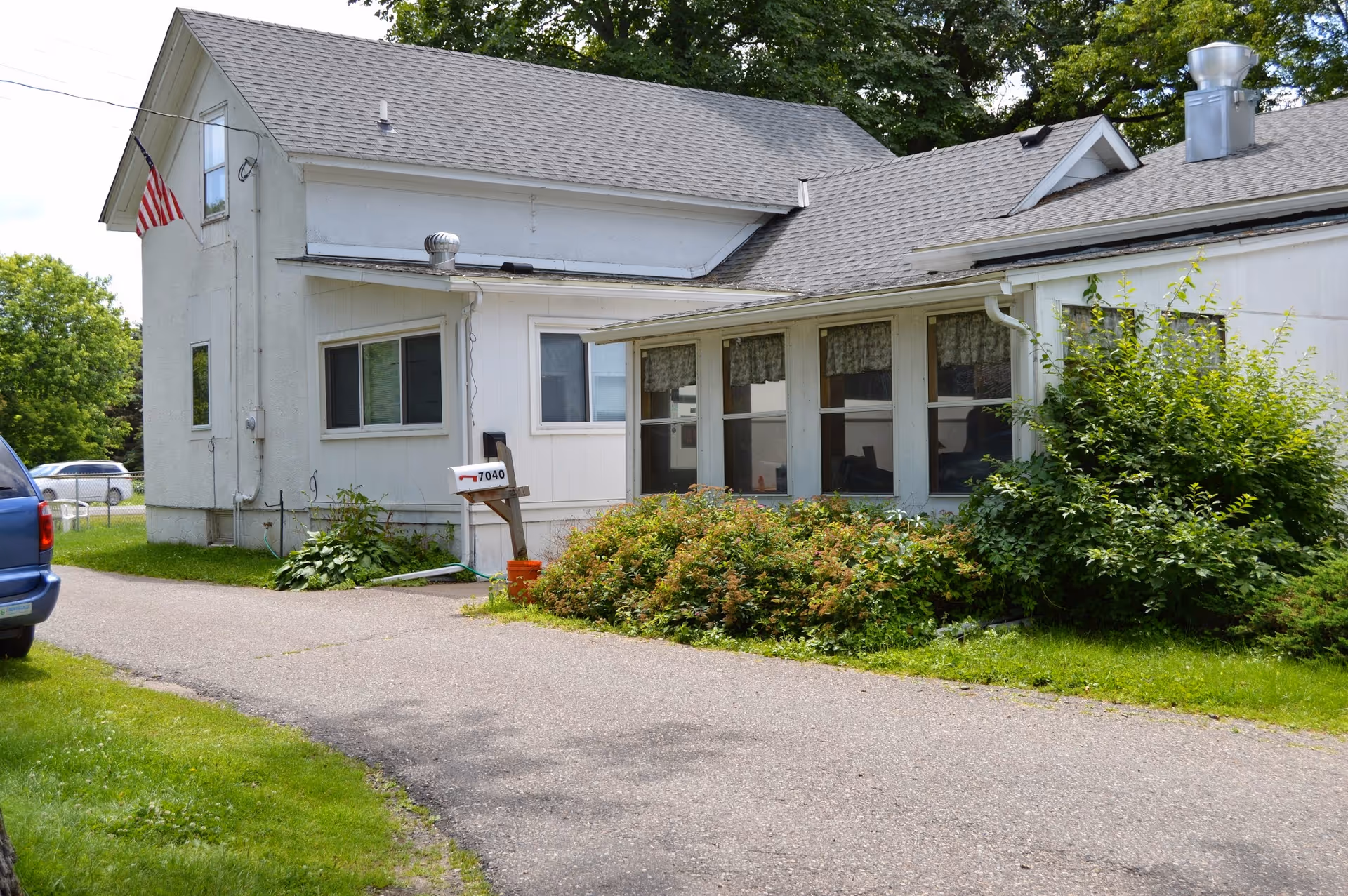 Exterior view of a white building with a gray roof, surrounded by greenery and bushes. There is a driveway leading up to the building, an American flag mounted on the side, and a mailbox with the number 7040. A blue vehicle is partially visible on the left side of the image.