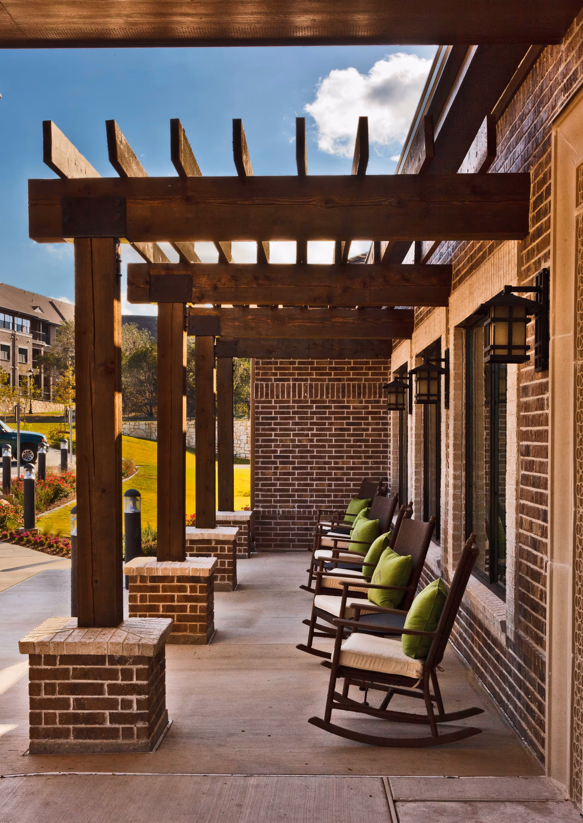 Outdoor covered patio area with wooden pergola beams and brick pillars. Several wooden rocking chairs with green cushions are lined up against a brick wall with windows and lantern-style wall lights. The area overlooks a landscaped garden with grass and flowers under a partly cloudy sky.