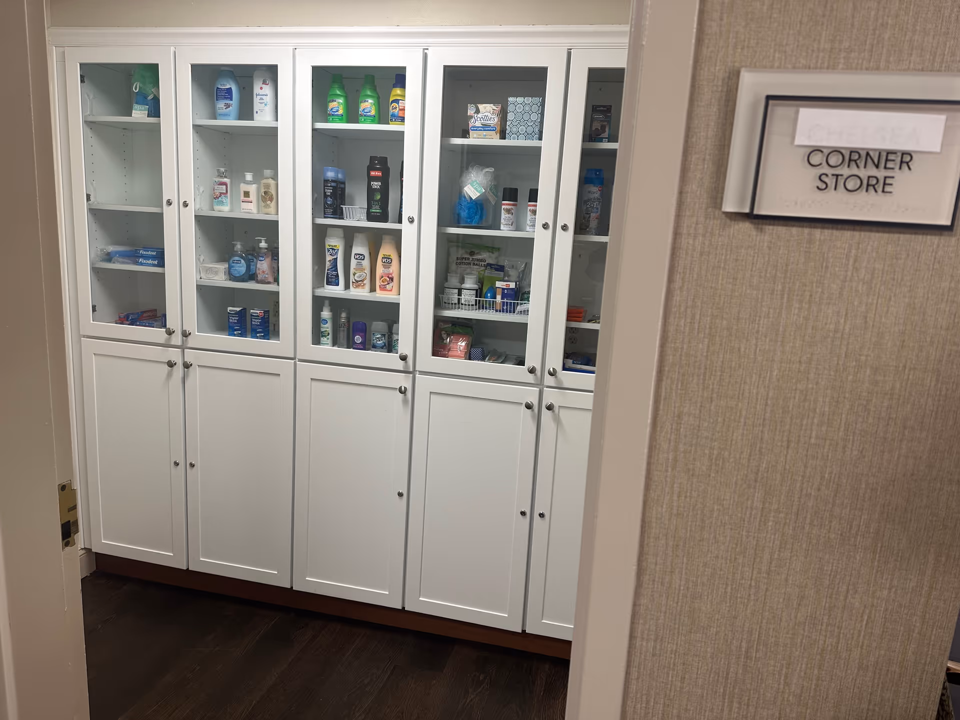 A white cabinet with glass doors displaying various personal care products such as lotions, shampoos, deodorants, and hygiene items. To the right of the cabinet, there is a wall sign that reads 'CORNER STORE' with a handwritten label above it.