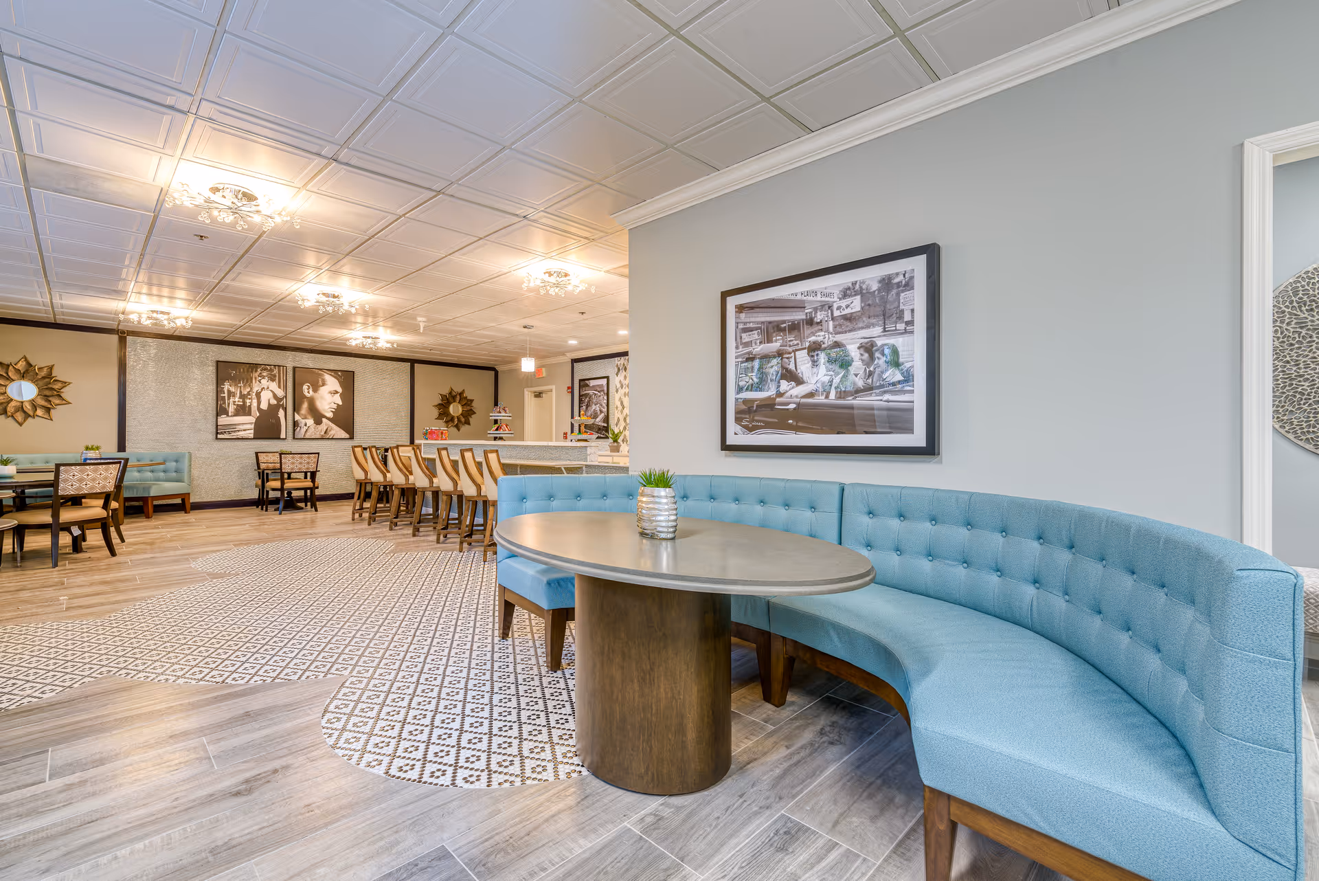 A spacious and well-lit dining area in a senior living facility featuring a curved blue upholstered bench around an oval table with a small plant centerpiece. The room has patterned tile flooring blending into wood-like tiles, multiple dining tables and chairs, decorative wall art including black and white photos, and elegant ceiling lights.