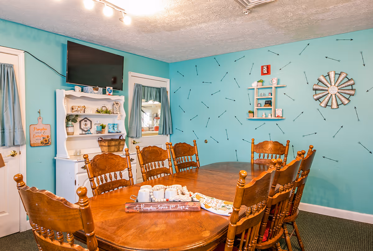 A cozy dining room with a wooden dining table surrounded by eight matching wooden chairs. The walls are painted turquoise with black arrow patterns. A white hutch with decorative items and a flat-screen TV mounted above it is against one wall. There are two doors with small windows and teal curtains. The room is warmly lit by ceiling track lights.