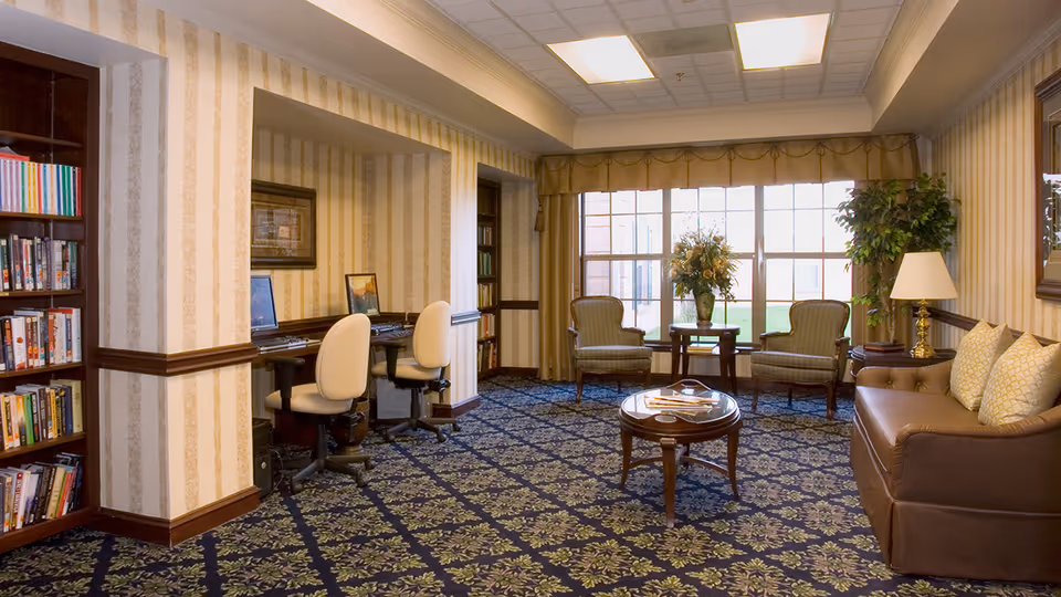 A cozy senior community lounge with a patterned carpet, a brown leather sofa with cushions, two armchairs around a small table with a flower arrangement, a side table with a lamp, and a built-in desk area with two office chairs and computers. Large windows with curtains allow natural light to fill the room. Bookshelves line the walls.