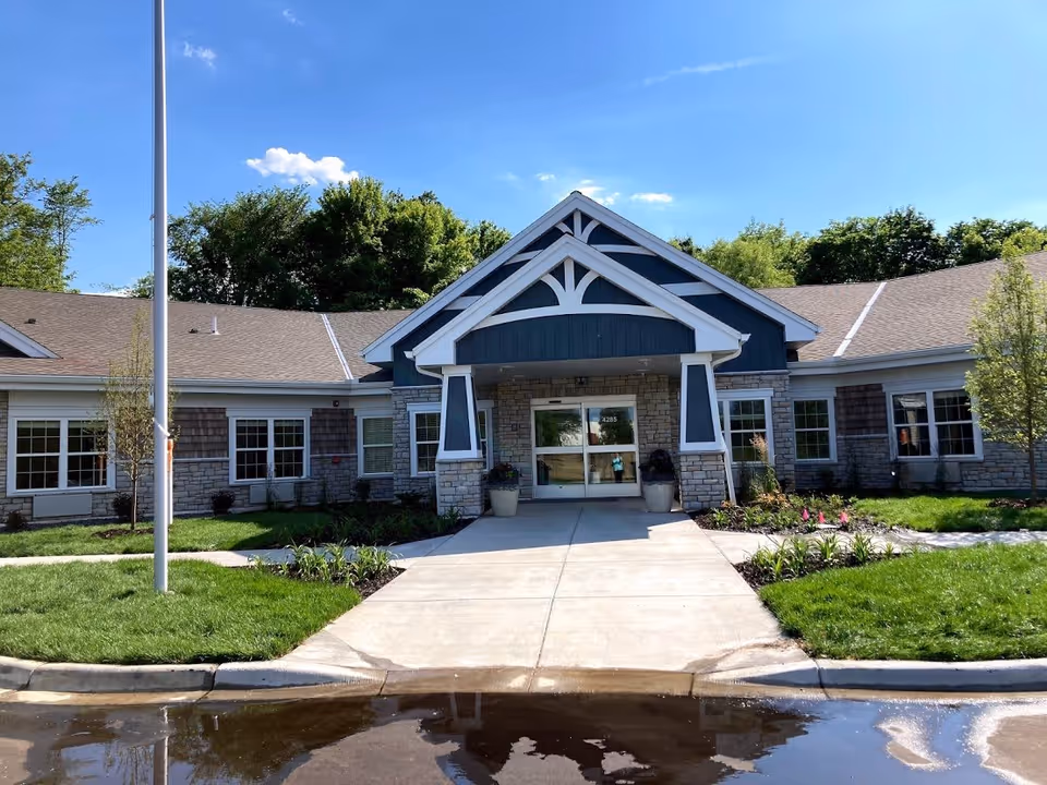 Front exterior view of NorBella Senior Living - Prior Lake building with a peaked roof entrance, stone and wood siding, large windows, a concrete walkway, green grass, small trees, and a clear blue sky.