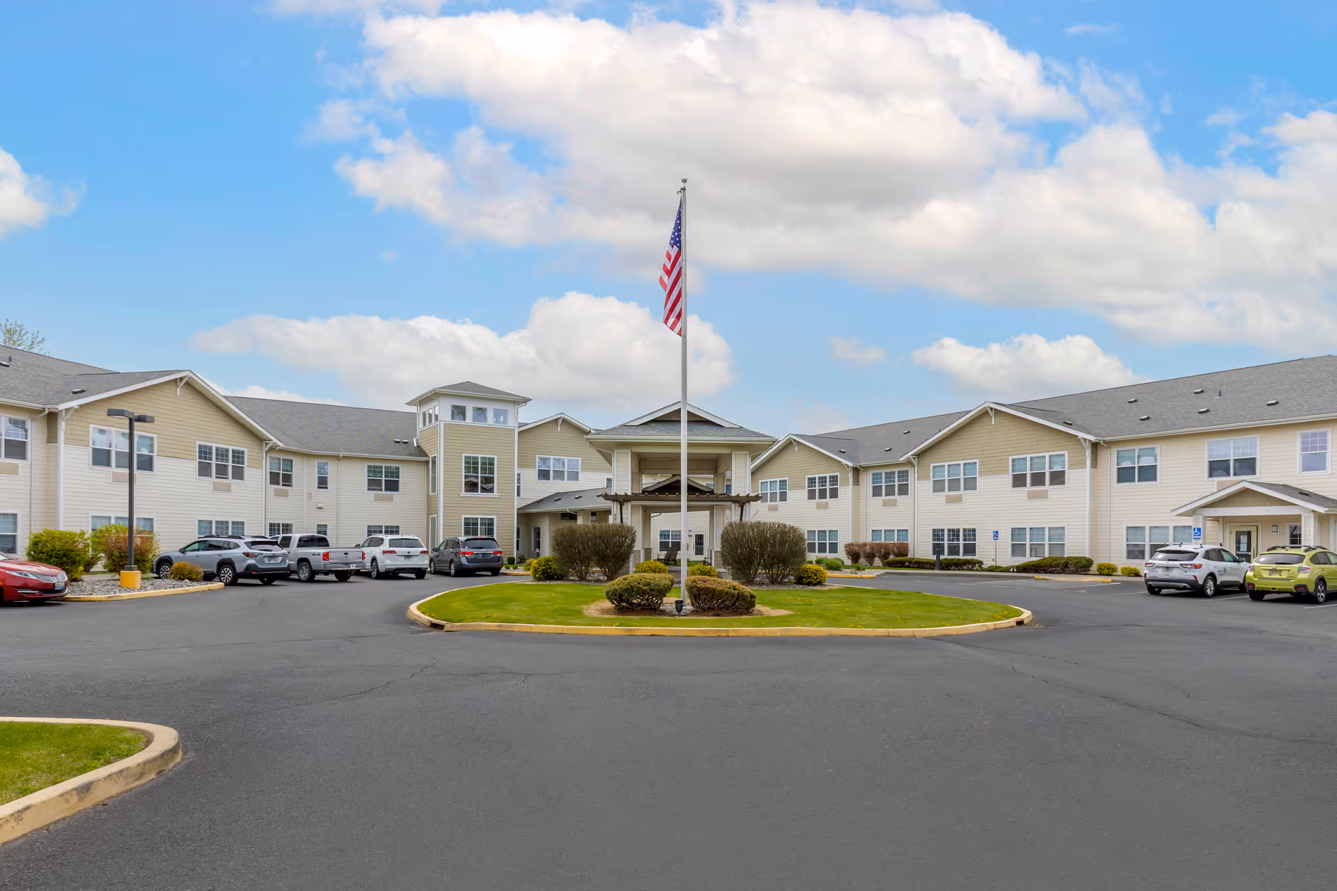 Front exterior of a multi-story senior living building with a circular driveway, parked cars, and an American flag in front.