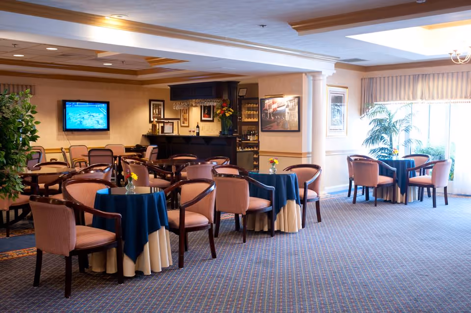 A spacious dining area with multiple round tables covered with blue and beige tablecloths, each surrounded by four cushioned chairs. The room has carpeted floors, a large window with curtains letting in natural light, framed artwork on the walls, a mounted flat-screen TV, and a small bar area with bottles and flowers.