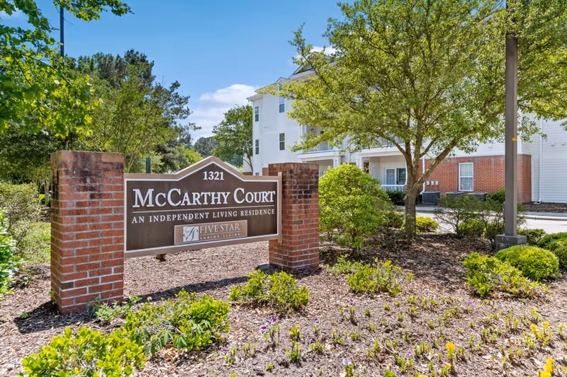 Outdoor view of the entrance sign for McCarthy Court, an independent living residence, surrounded by landscaped greenery, trees, and a multi-story building in the background under a blue sky.