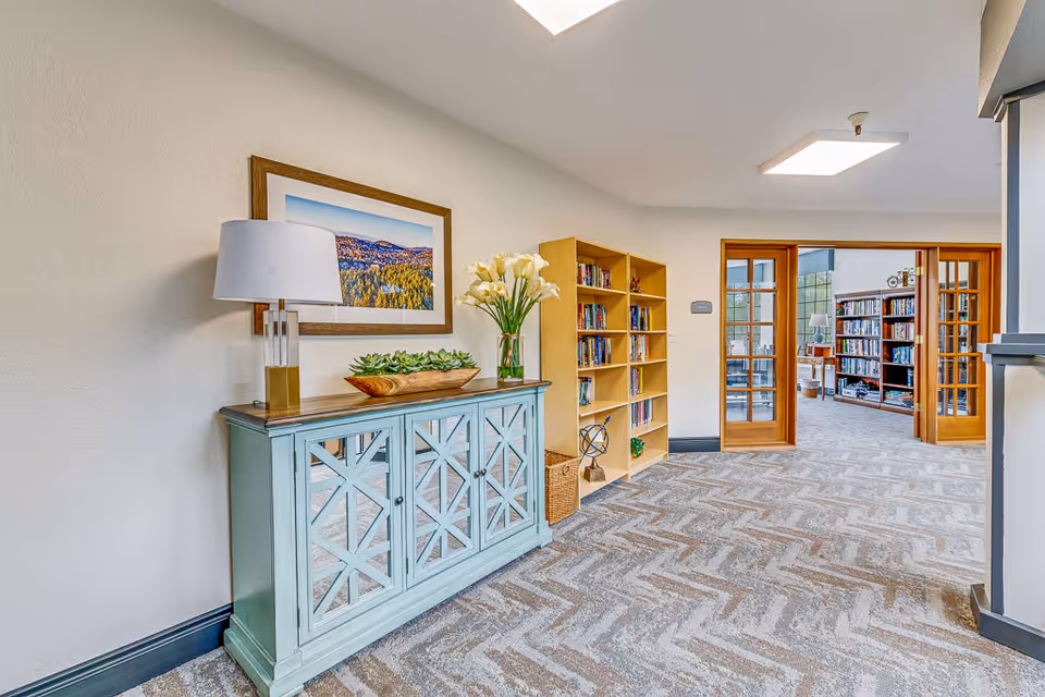 A hallway in a senior living facility featuring a light blue cabinet with a lamp, a wooden bowl with succulents, and a vase of white flowers on top. Above the cabinet is a framed landscape photograph. To the right, there is a wooden bookshelf filled with books and a wicker basket underneath. The hallway leads to a room with glass-paned wooden double doors, revealing more bookshelves and seating inside. The floor is carpeted with a geometric pattern.