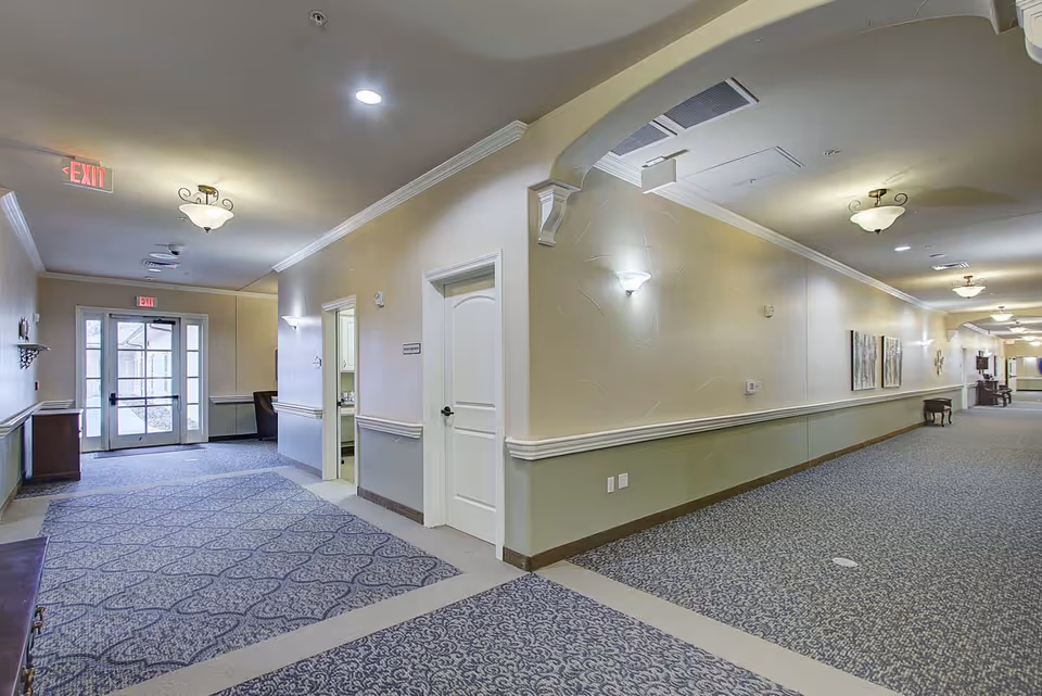 Interior view of a well-lit hallway in a senior living facility with patterned carpet, beige walls with white trim, wall sconces, ceiling lights, exit signs, and doors leading to rooms. There are some pieces of furniture and artwork along the hallway.