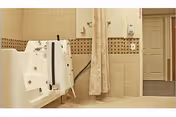 A bathroom with a walk-in bathtub featuring safety handles and a handheld showerhead. The walls are tiled with beige tiles and a decorative brown tile border. There is a privacy curtain hanging from a rod, and a doorway leading to another room is visible in the background.