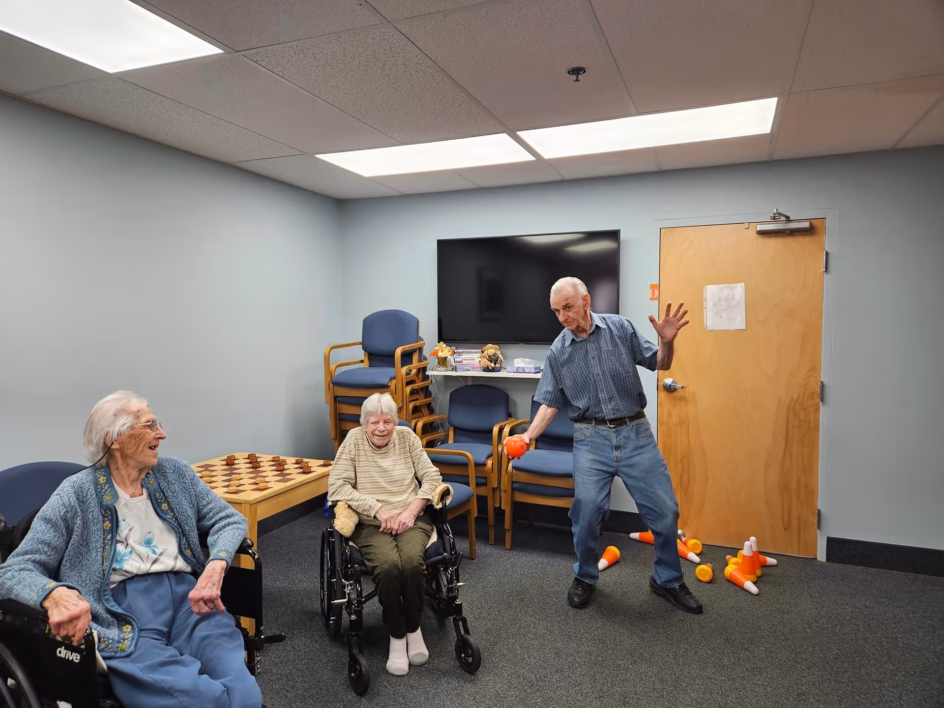 Three elderly individuals in a room at Manatawny Manor. Two women are seated, one in a wheelchair and the other in a chair, smiling and watching a man who is standing and appears to be playing a game with orange and white plastic pins and balls. The room has a TV mounted on the wall, stacked chairs, a wooden door, and a table with a checkerboard pattern.