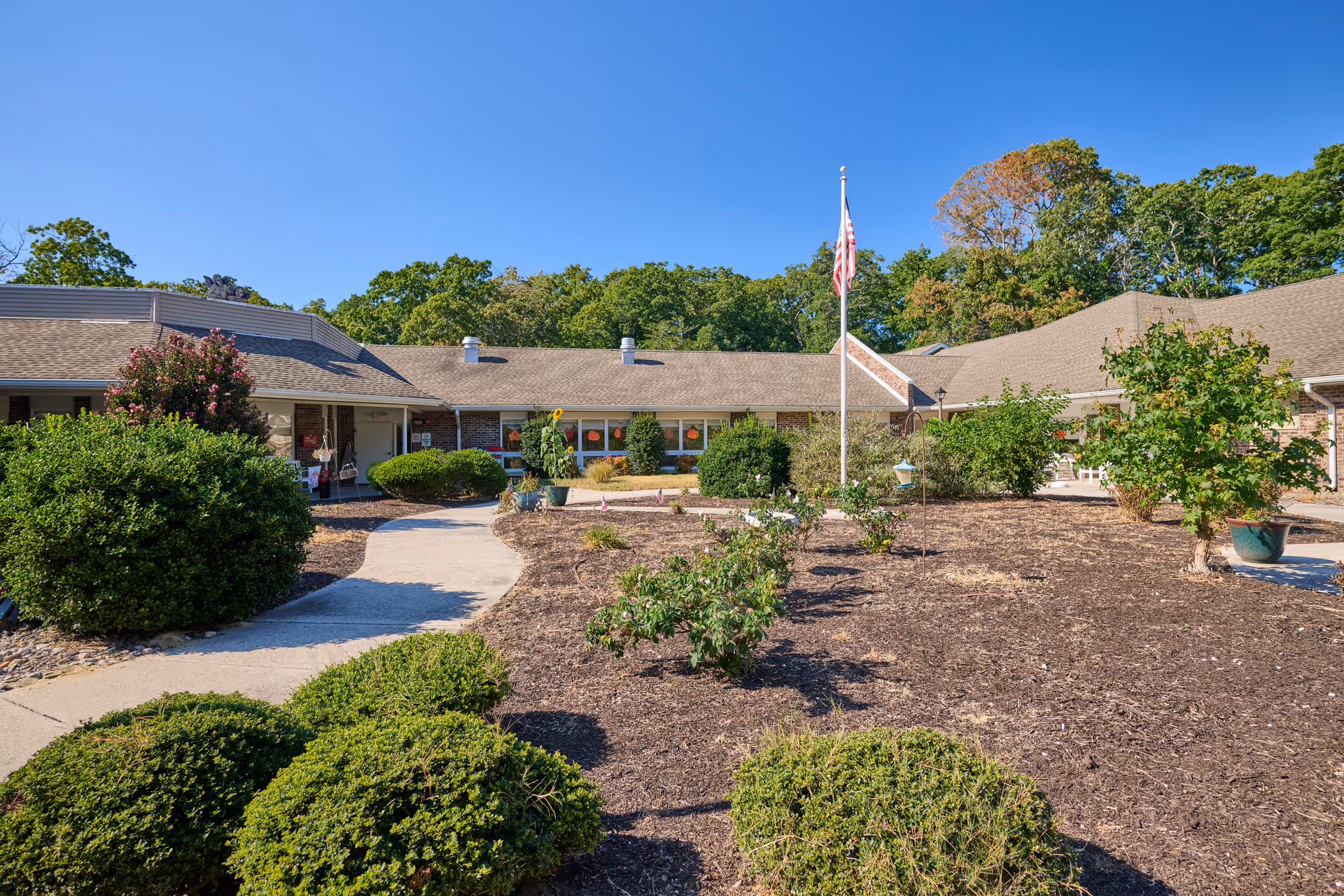 Exterior view of Pelican Pointe Post Acute Nursing & Rehabilitation facility showing a single-story building with a beige roof and brick walls. There is a paved walkway leading to the entrance, surrounded by landscaped bushes and small trees. An American flag is raised on a flagpole in the garden area under a clear blue sky.