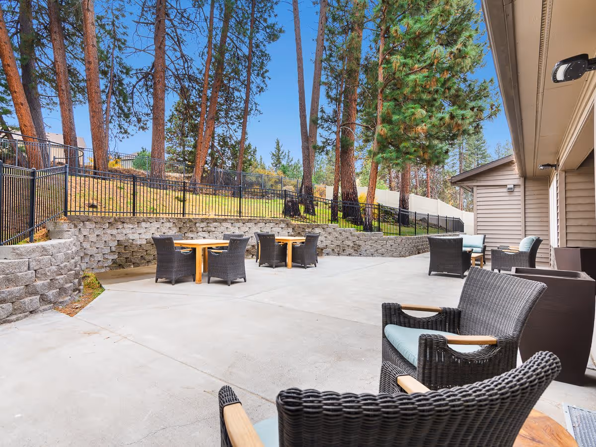 Outdoor patio area at Mill View Memory Care with several wicker chairs and wooden tables on a concrete surface, surrounded by a retaining wall and tall pine trees under a clear blue sky.