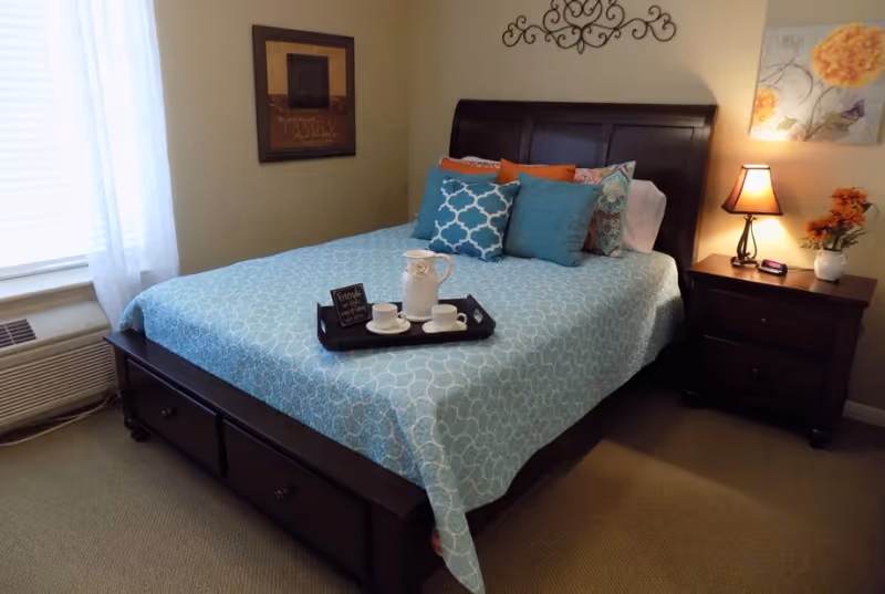 Sunlit bedroom with a wooden bed dressed in turquoise patterned bedding, decorative pillows, a tray on the bed, and a nightstand with a lamp.