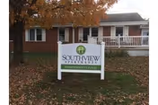 A white sign in front of a brick building with a porch and white railing. The sign reads 'Southview Apartments Assisted Living' and is placed on a grassy area with fallen autumn leaves. A tree with orange leaves is visible to the left of the sign.