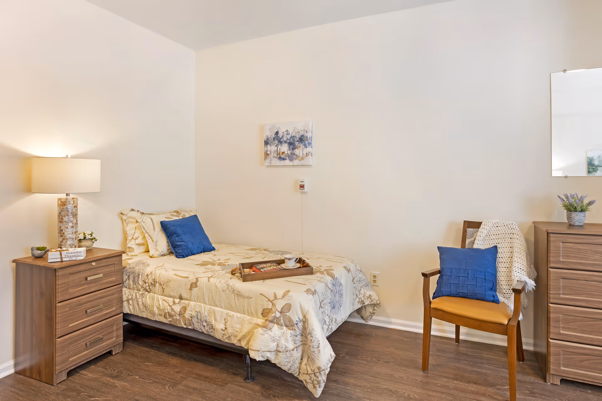 A cozy bedroom in a senior living facility with a single bed covered in a floral bedspread and a blue pillow. Next to the bed is a wooden nightstand with a lamp, a small plant, and a book. There is a wooden chair with a blue cushion and a white knitted throw blanket. A wooden dresser with a small potted plant and a mirror above it is also visible. The walls are light-colored and the floor is wooden.
