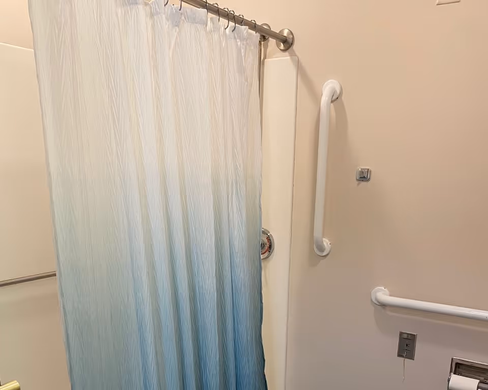 Bathroom with a shower area featuring a white and blue gradient shower curtain. There are white grab bars mounted on the wall for support and safety. The walls are painted a light beige color.
