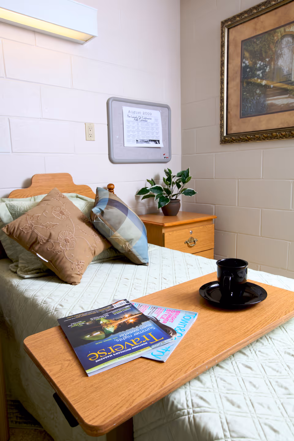 A neatly made single bed with decorative pillows in a room with light-colored walls. A wooden bedside table holds a small potted plant. A wooden overbed table is placed on the bed, holding a black cup and saucer and two magazines. A framed picture hangs on the wall above the bedside table.