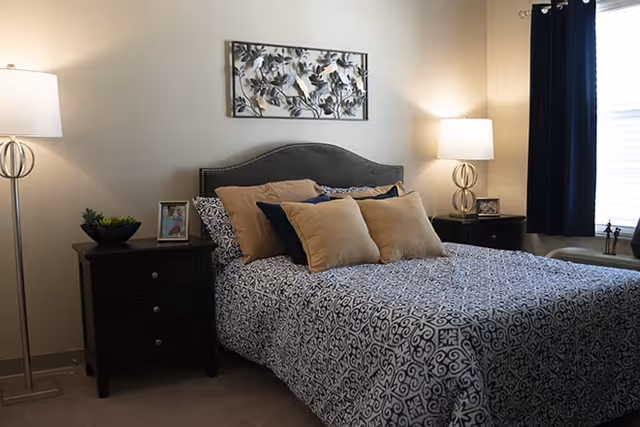 A neatly made bed with patterned blue and white bedding and several beige and navy pillows in a bedroom. There are two dark wooden nightstands on either side of the bed, each with a lamp. A decorative metal wall art piece hangs above the bed, and a window with dark curtains is visible on the right side.