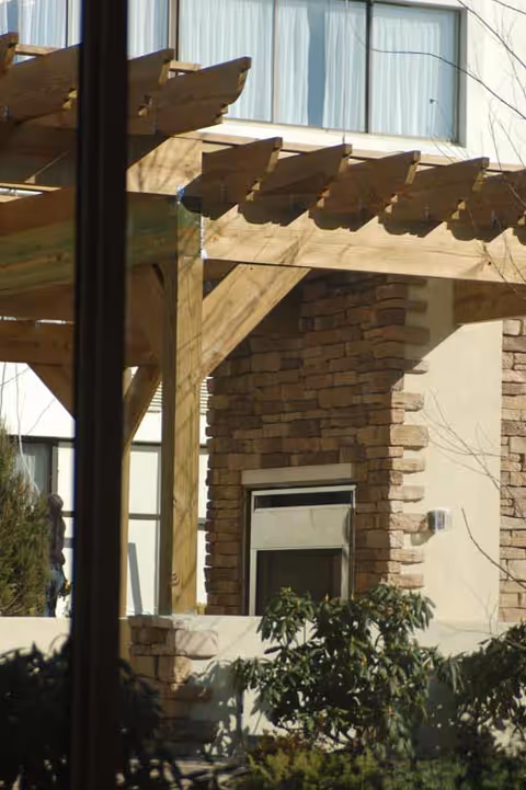 Wooden pergola over a stone-clad exterior wall with windows and landscaping at a senior living facility.