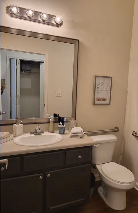 A bathroom with a white sink set in a beige countertop with dark wooden cabinets underneath. Above the sink is a large rectangular mirror with a silver frame and a light fixture with four bulbs. To the right of the sink is a white toilet with a grab bar mounted on the wall behind it. A small framed picture with the word 'BLESSED' hangs on the wall above the toilet. Various toiletries and folded towels are placed on the countertop.
