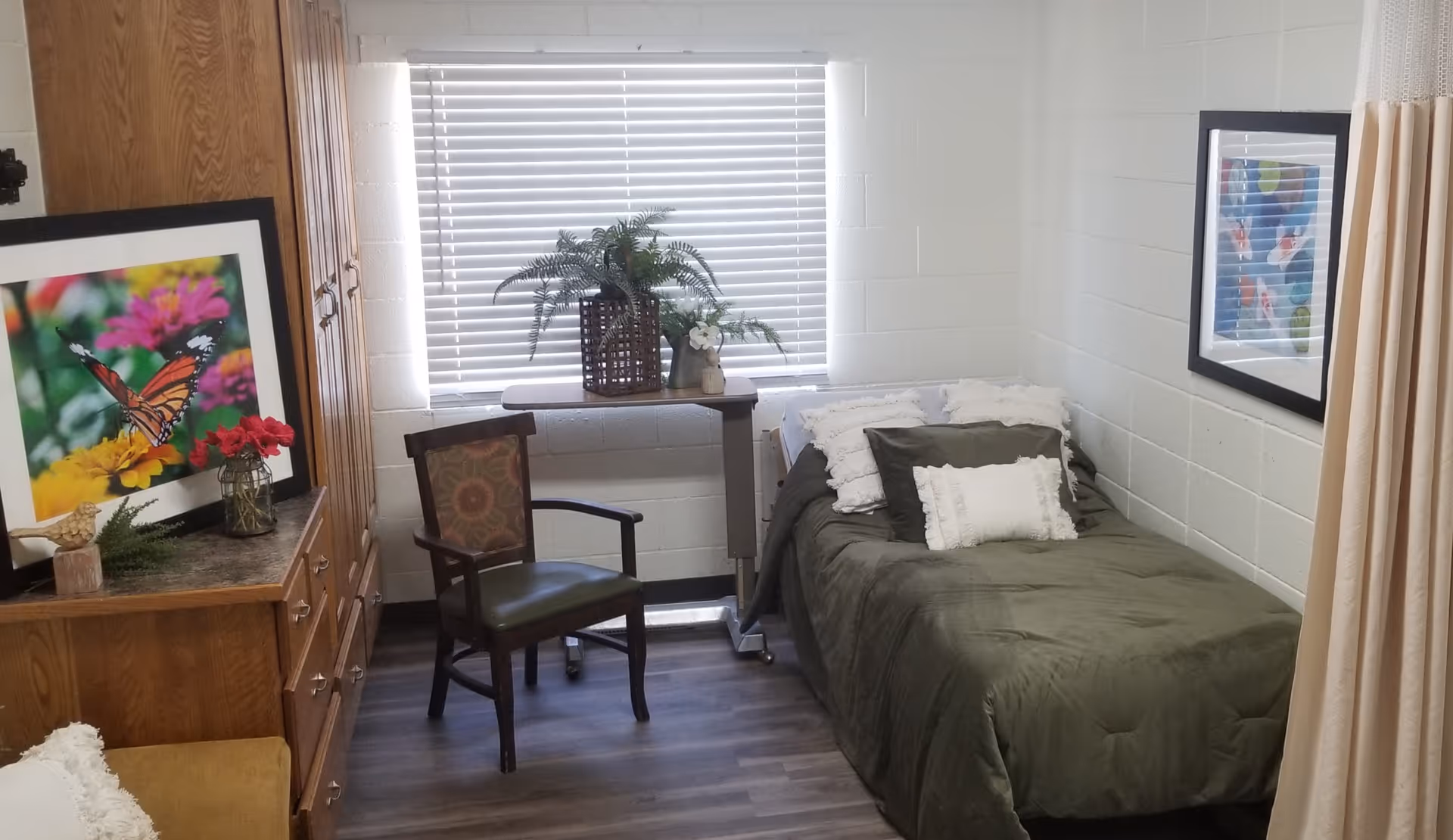 A small bedroom with a single bed covered in a green comforter and white pillows. There is a wooden chair with patterned upholstery next to a small table holding a plant in front of a window with closed blinds. On the left side, there is a wooden dresser with a framed butterfly picture and a small vase with red flowers. The walls are white and made of cinder blocks, and the floor has wood-like vinyl flooring.