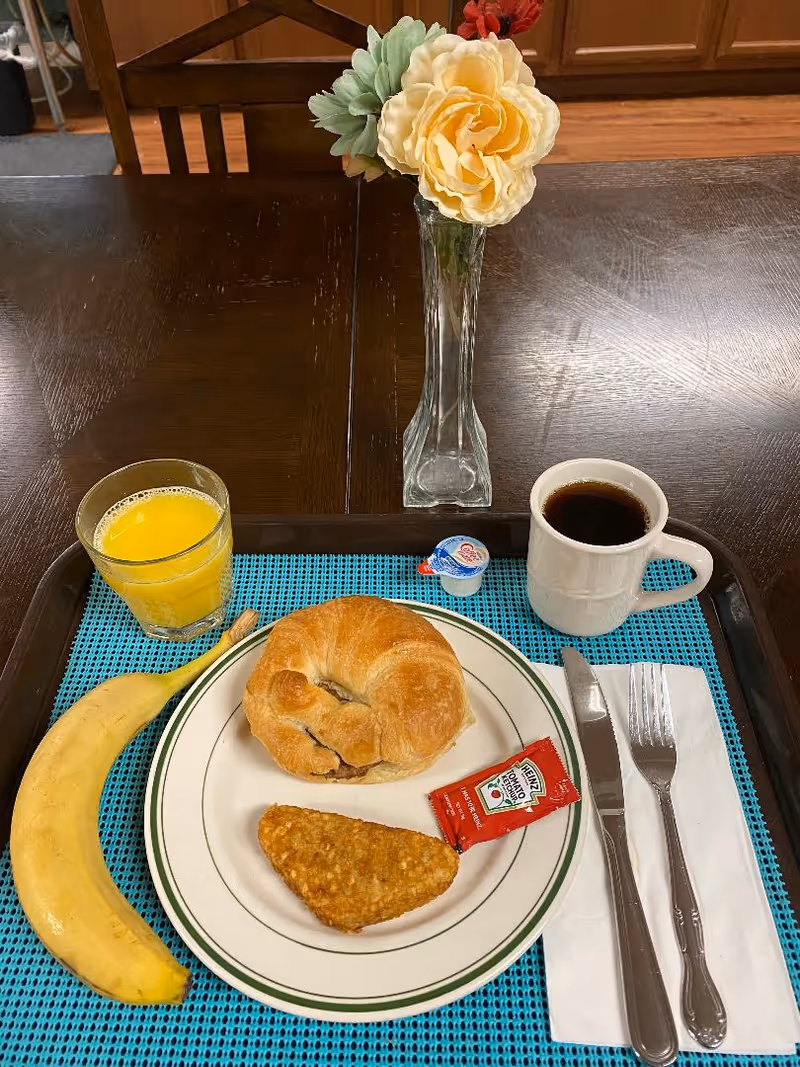 Breakfast tray on a table with a croissant, hash brown, banana, glass of orange juice, cup of coffee, utensils and a vase of flowers.