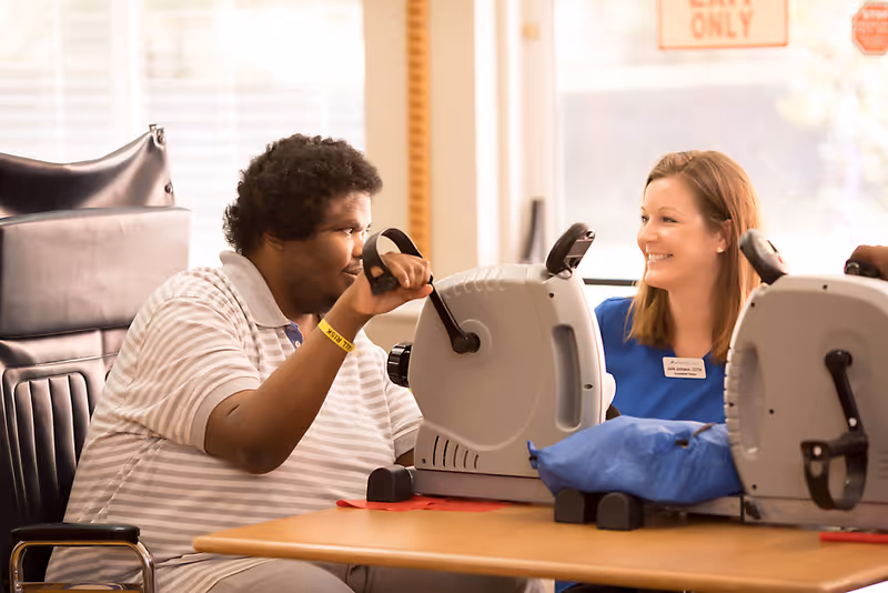 A person in a wheelchair using a hand pedal exerciser at a table, assisted by a smiling female therapist wearing a blue uniform and name tag, in a bright room with large windows.