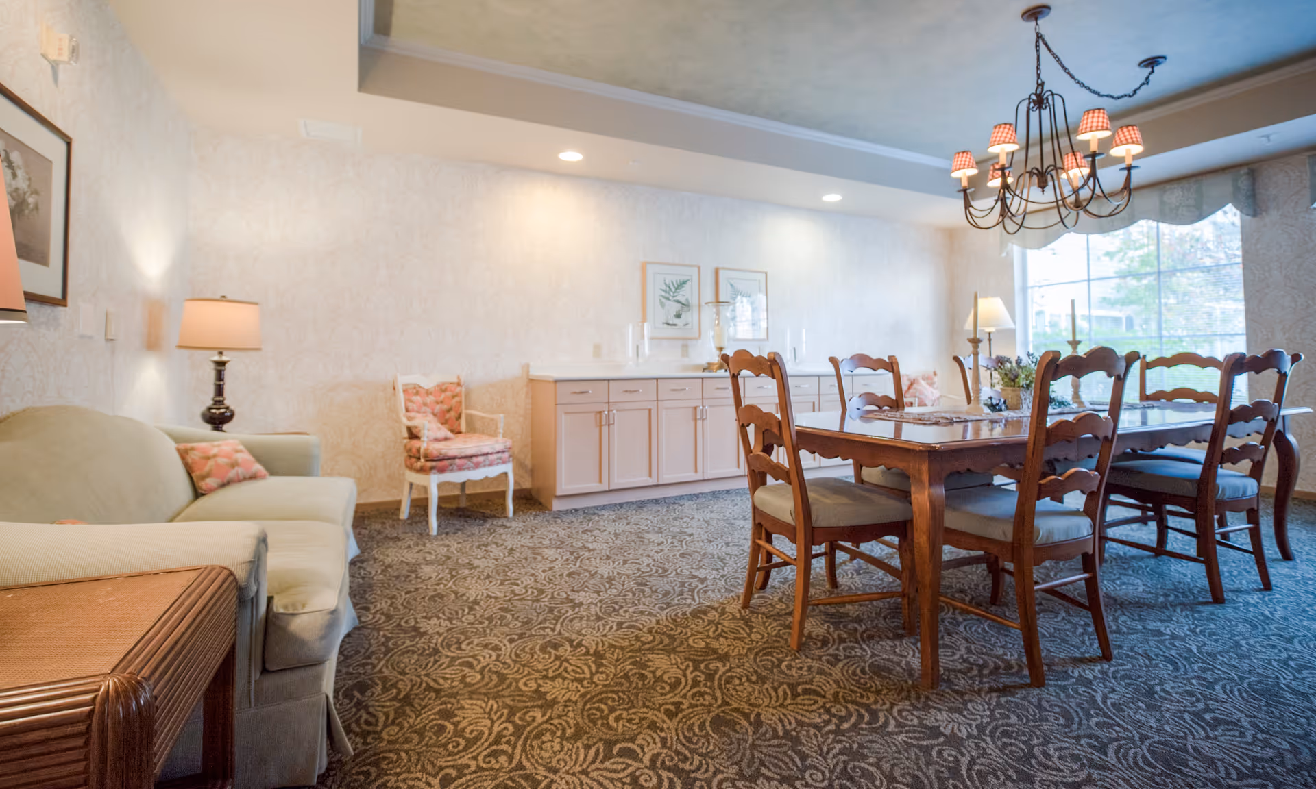 A bright communal dining room with a wooden table and chairs, a chandelier, sofa and sideboard by a large window.