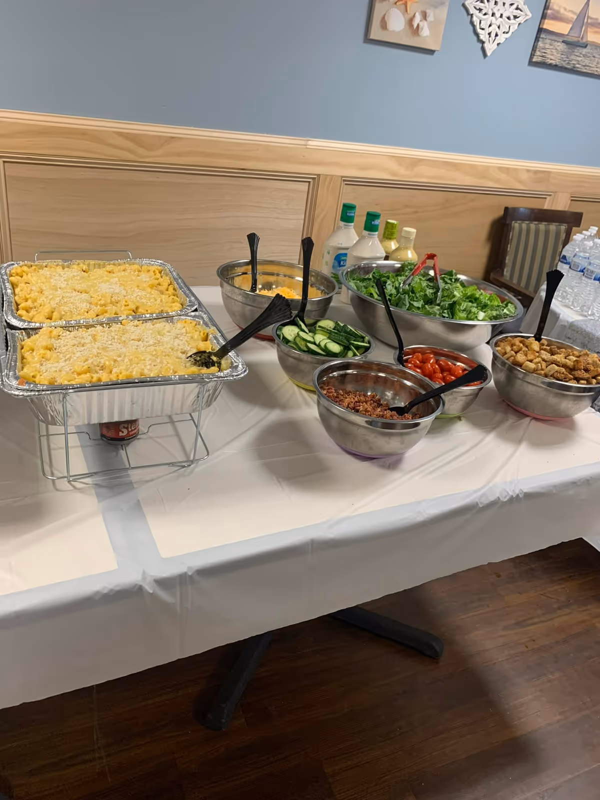 A buffet table with trays of macaroni and cheese, bowls of salad ingredients including lettuce, cucumbers, cherry tomatoes, bacon bits, croutons, and several bottles of salad dressing. Bottled water is visible in the background on a separate table.