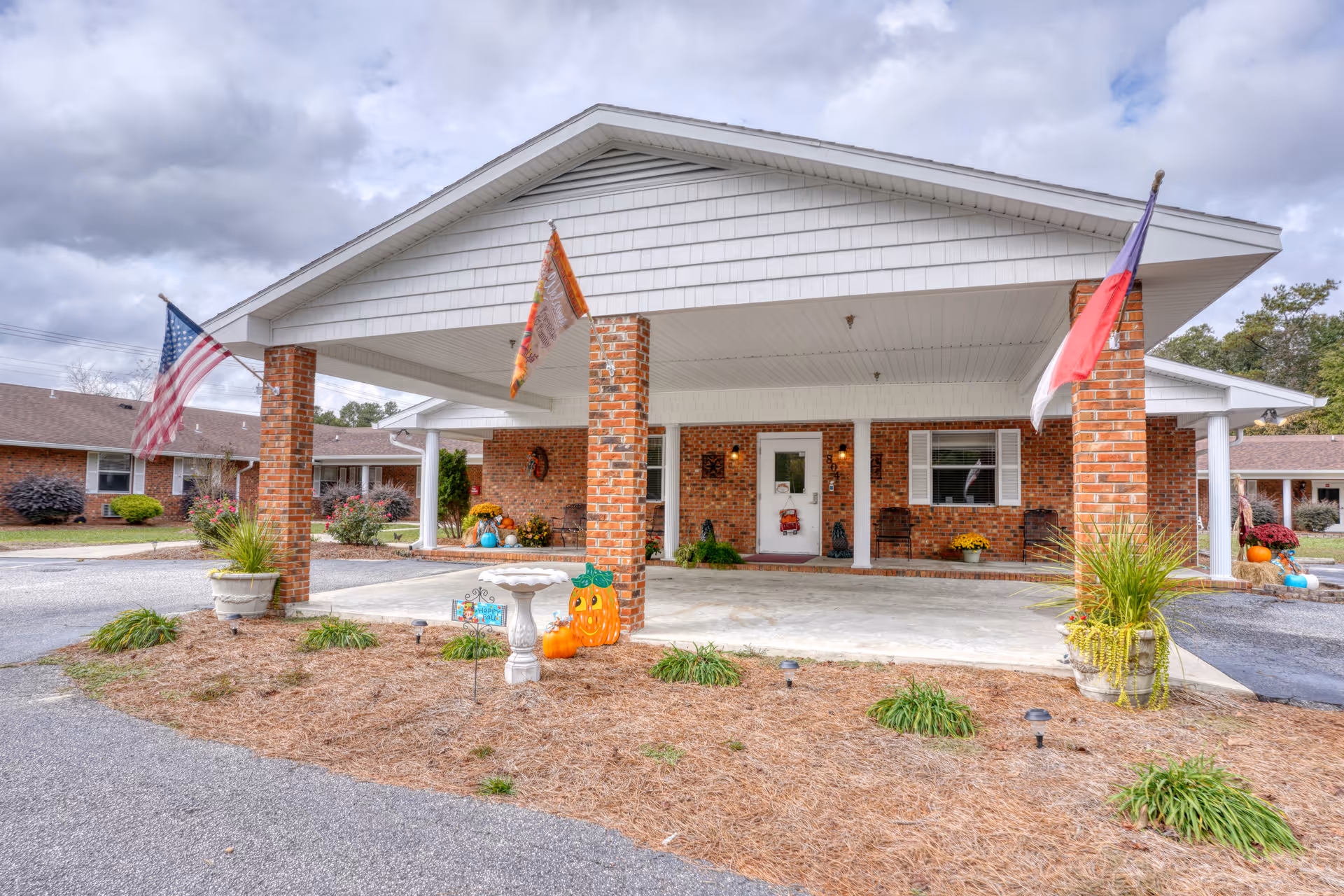 Front exterior view of a single-story brick building with a covered entrance supported by brick columns. The entrance area is decorated with autumn-themed items including pumpkins and a birdbath. There are three flags displayed: an American flag, a flag with orange and white colors, and a red, white, and blue flag. The sky is cloudy and there are some shrubs and plants around the building.
