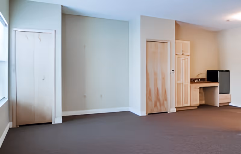 Empty studio-style living area with carpet, two wooden closet doors, a small kitchenette/desk and a mini refrigerator.
