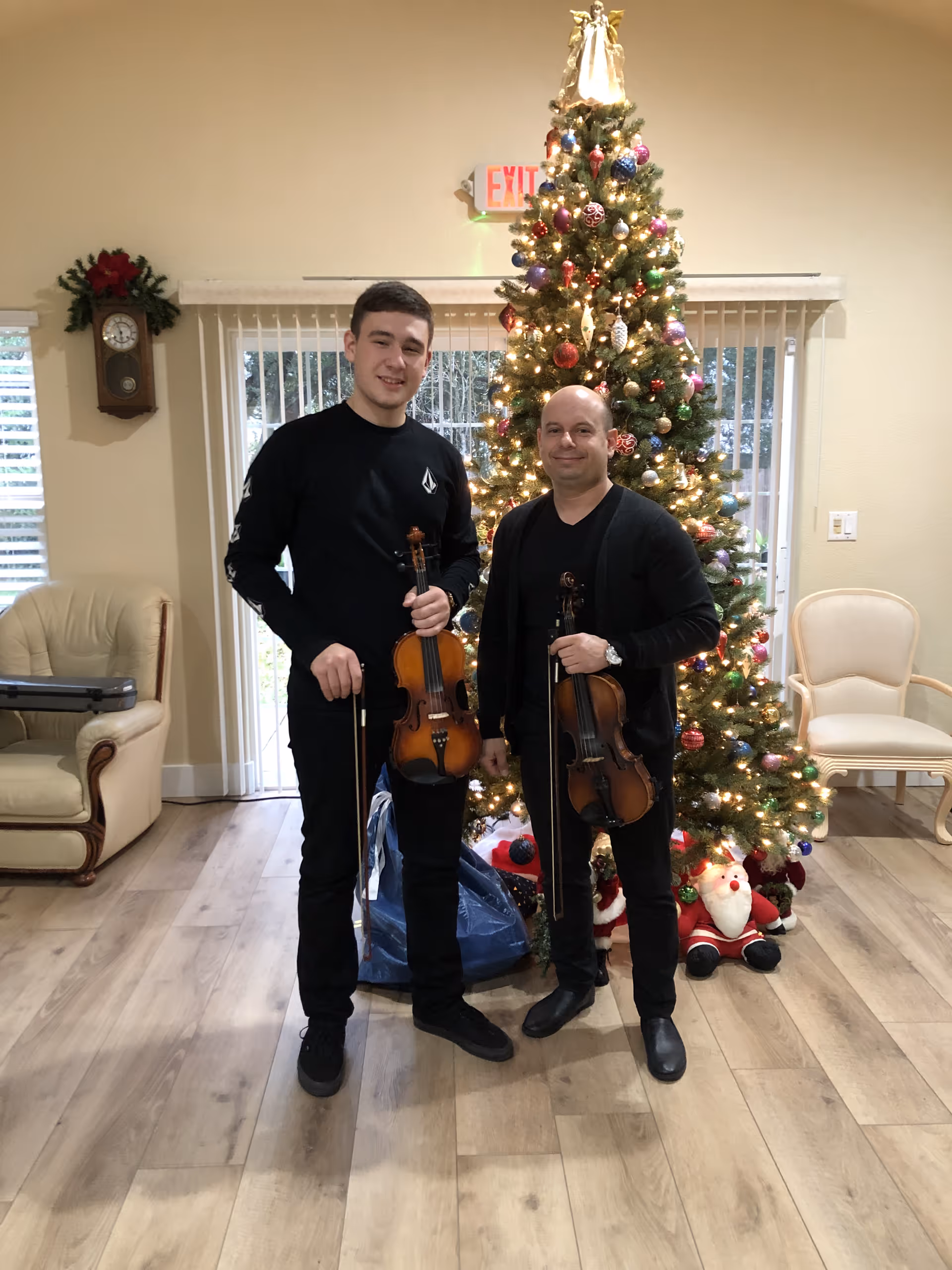 Two men holding violins stand in front of a decorated Christmas tree in a communal living room.