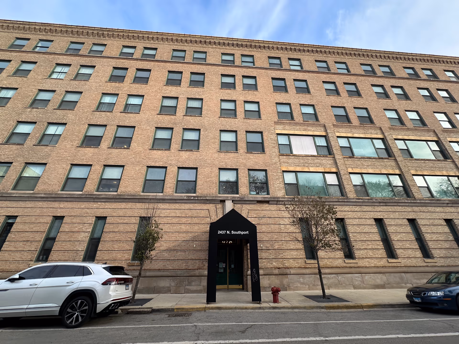 Exterior view of a multi-story brick building with many windows, a black awning over the entrance displaying the address 2437 N. Southport, two small trees on the sidewalk, and parked cars along the street.