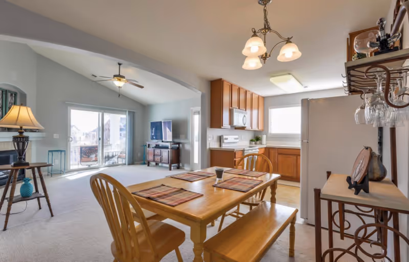 A bright and spacious open-plan living area in a senior living facility featuring a wooden dining table with four chairs and placemats, a kitchen with wooden cabinets, white appliances including a refrigerator and stove, and a living room area with a ceiling fan, a TV on a stand, and sliding glass doors leading to an outdoor balcony.