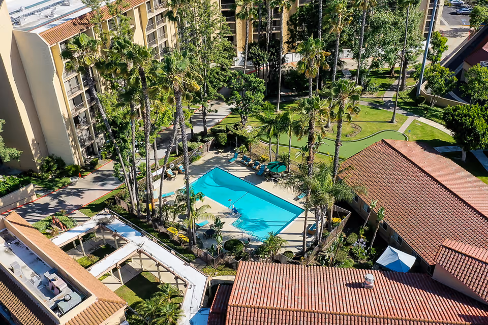 Aerial view of an outdoor area at a senior living facility featuring a rectangular swimming pool surrounded by lounge chairs and umbrellas, tall palm trees, green lawns, and several buildings with red-tiled roofs.